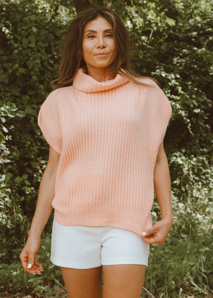 Woman wearing a pink sleeveless sweater and white shorts standing in a natural setting.