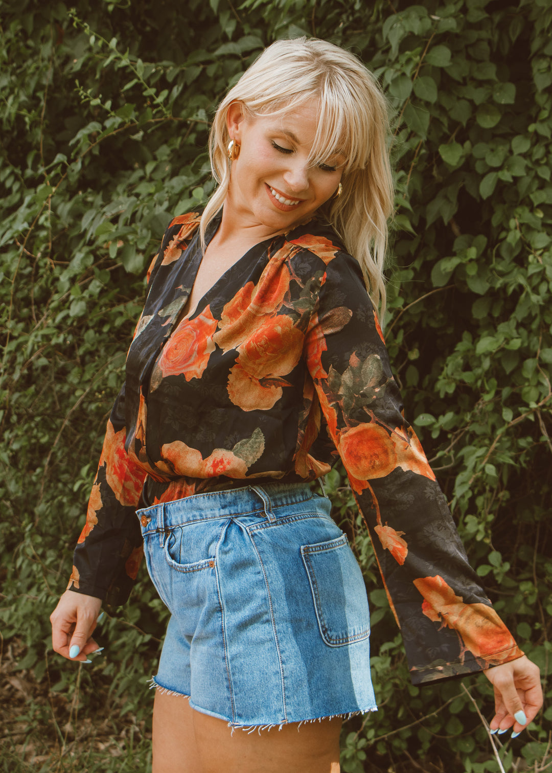 Woman wearing a floral blouse and denim shorts standing in front of green foliage