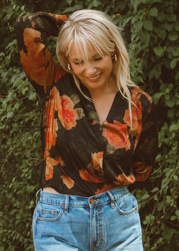 Woman wearing a floral blouse and jeans standing in front of green foliage
