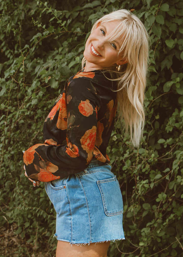 Woman wearing a floral blouse and denim shorts standing against a green leafy background