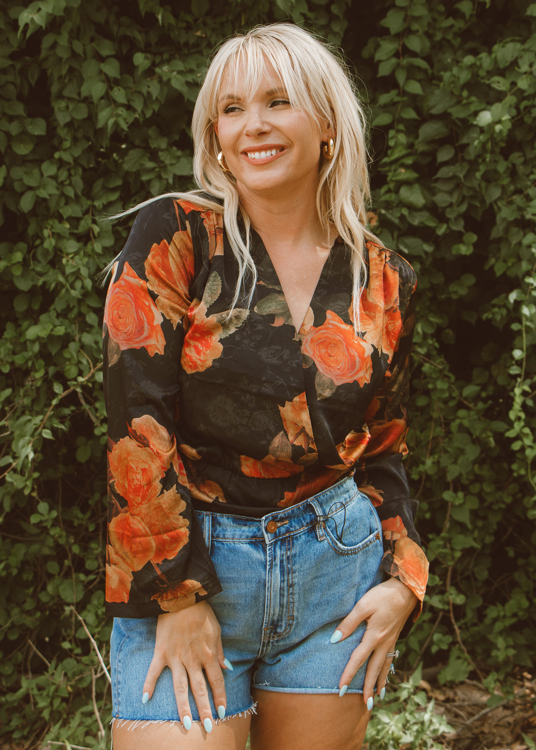 Woman wearing a floral blouse and denim shorts standing against a green leafy background