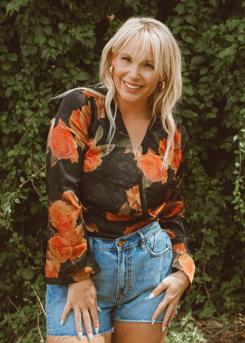 Woman wearing a floral blouse and denim shorts standing in front of green foliage
