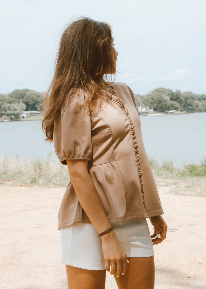 Woman standing by a lake wearing a beige blouse and white shorts.