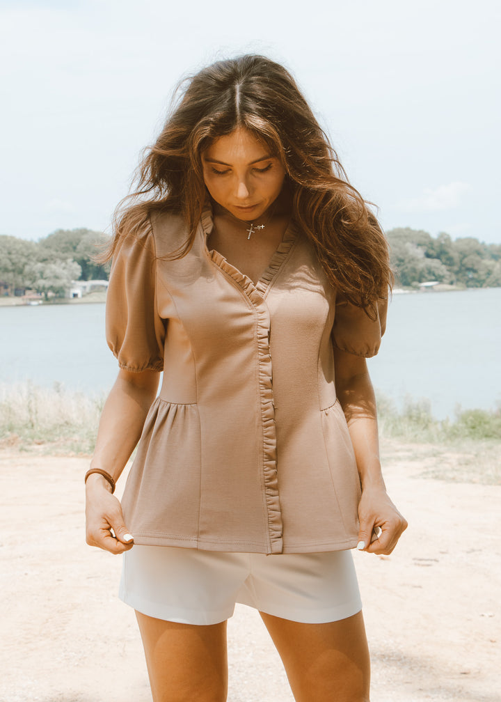 Woman wearing a beige top and white shorts standing by a lake.