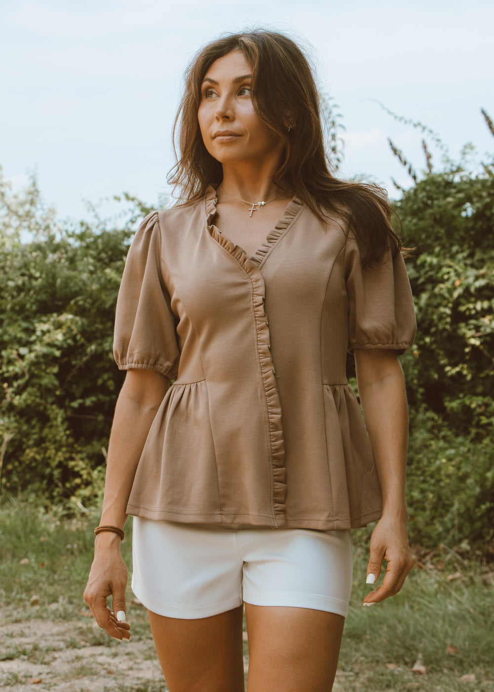 Woman wearing a brown blouse and white shorts outdoors with greenery in the background