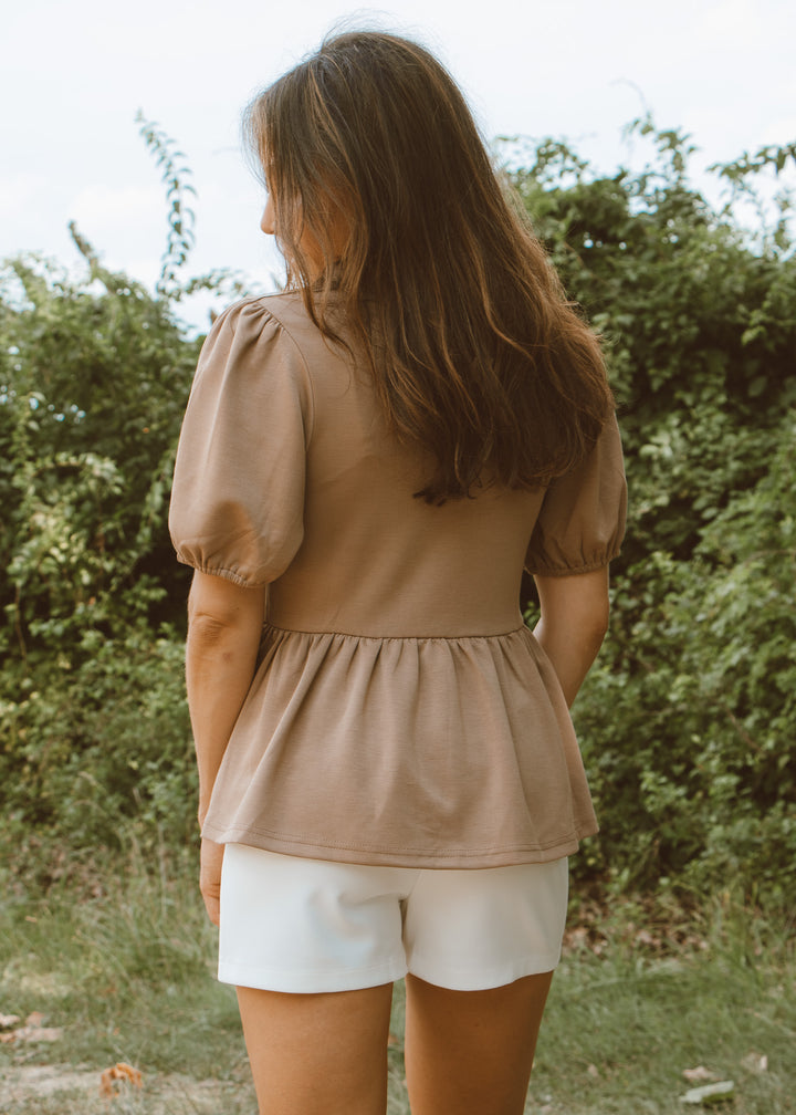 Woman wearing a beige top and white shorts standing in a natural setting with greenery.