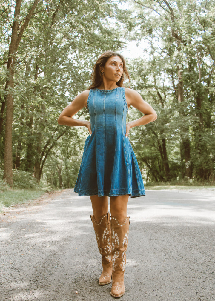 Woman in a blue dress and cowboy boots standing on a road with trees in the background