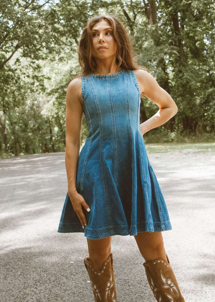 Woman wearing a blue denim dress and brown boots standing outdoors with trees in the background