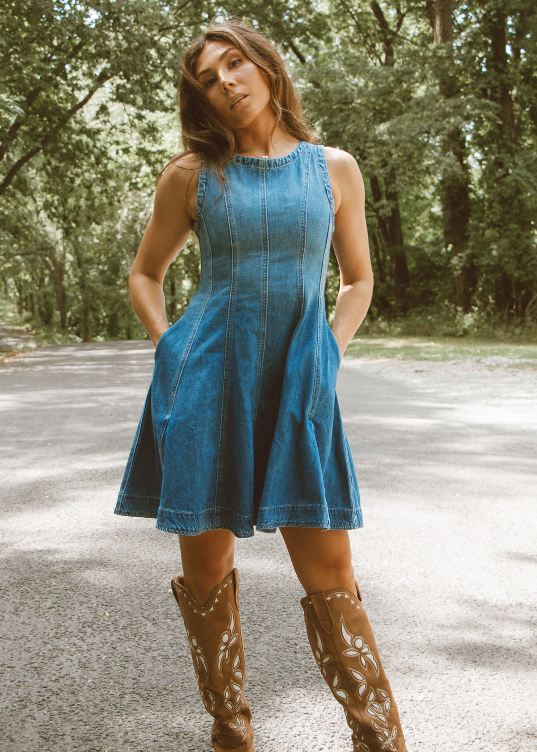 Woman wearing a blue dress and brown boots standing on a road with trees in the background