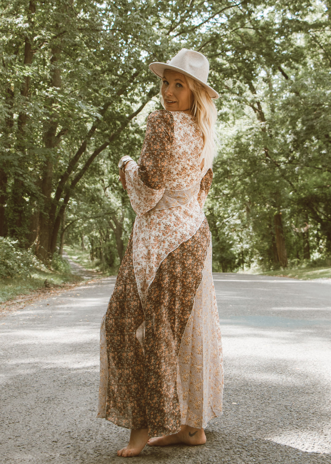 Woman in a floral dress and hat standing on a road surrounded by trees