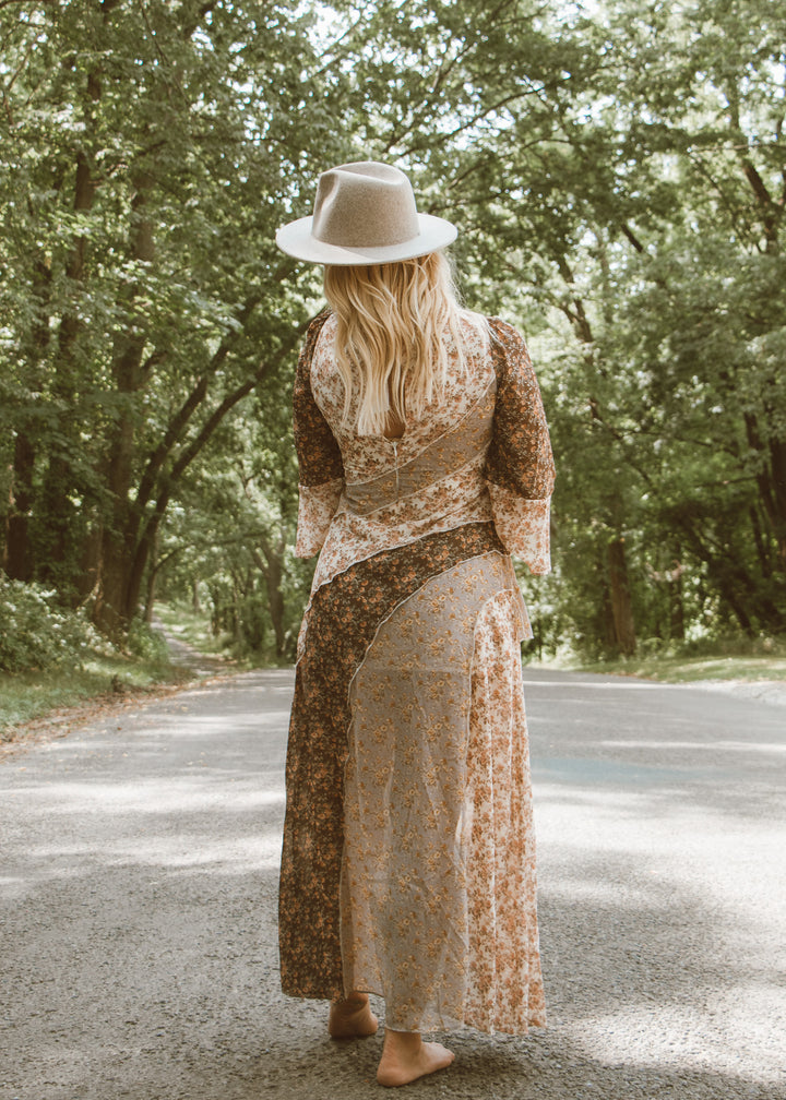 Woman in a floral dress and hat standing on a road surrounded by trees