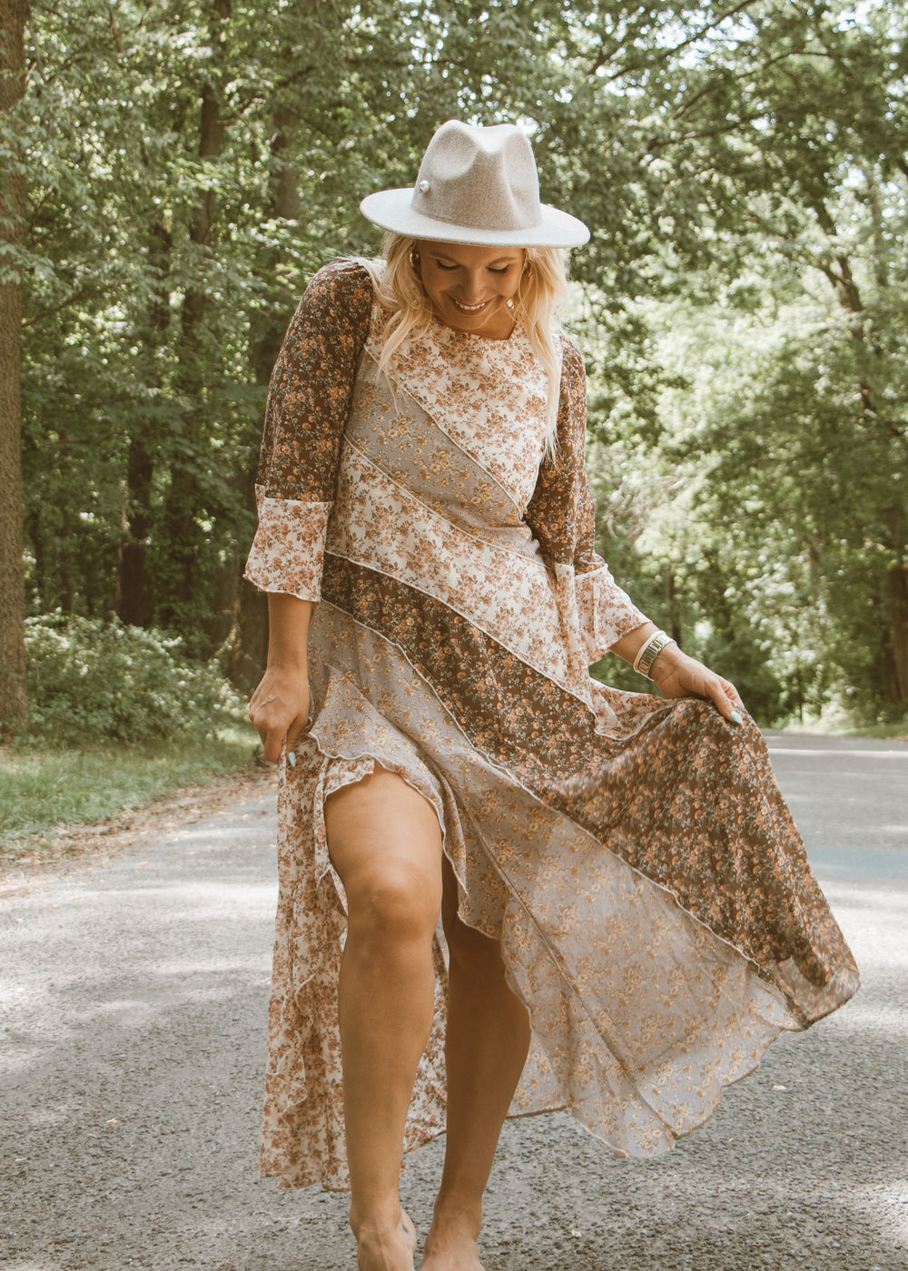 Woman in a floral dress and hat standing on a road with trees in the background