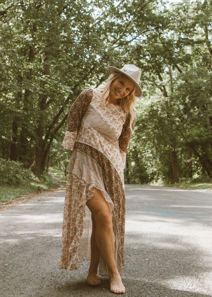 Woman in a floral dress and hat standing on a road surrounded by trees