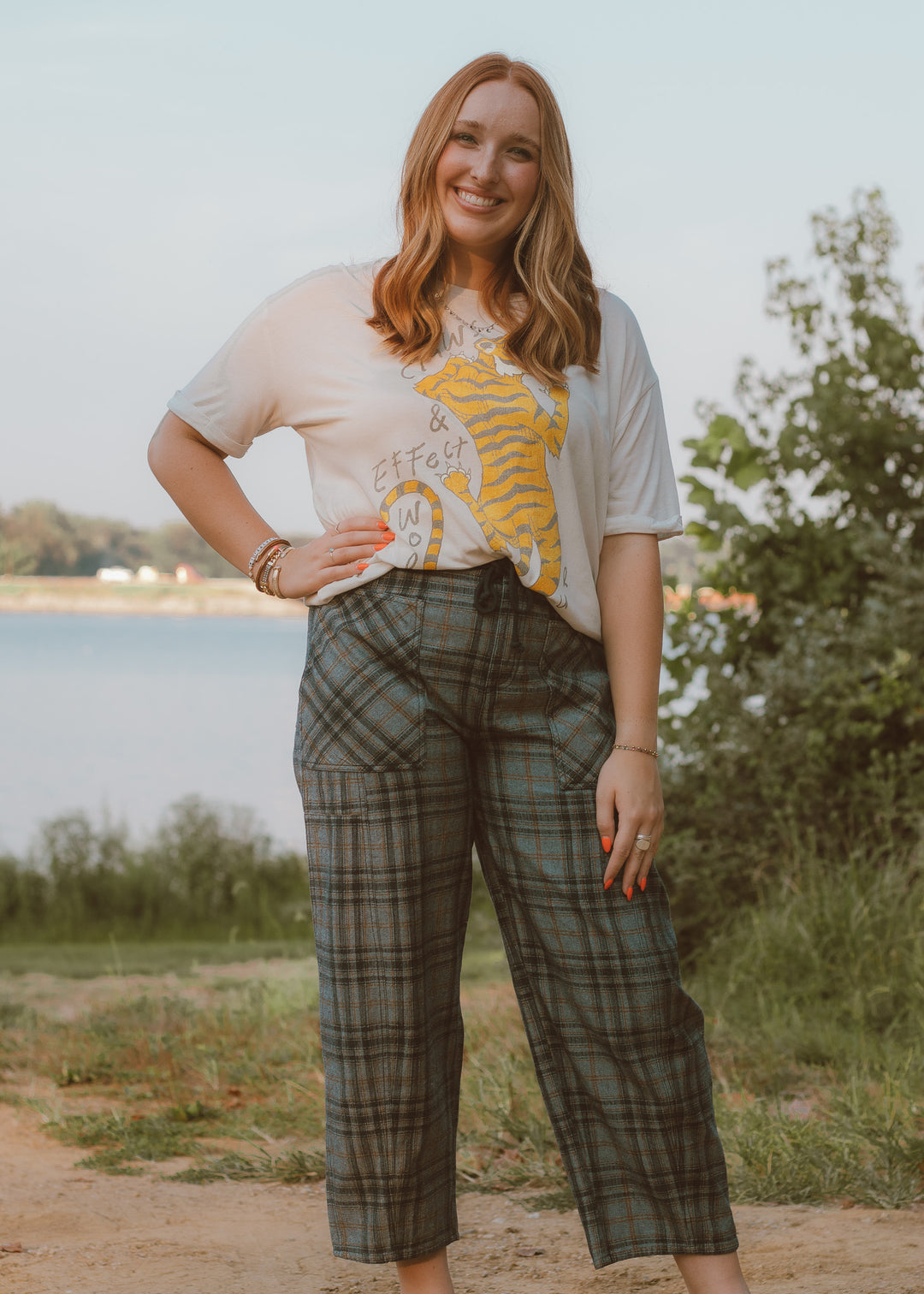 Woman wearing a graphic t-shirt and plaid pants standing outdoors near a body of water.