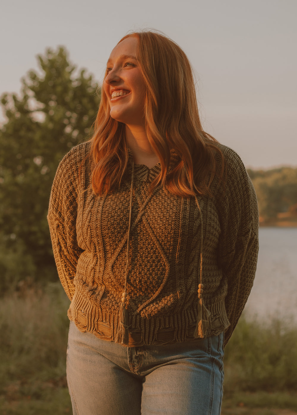 Woman wearing a green patterned sweater standing by a lake with a warm, golden light.