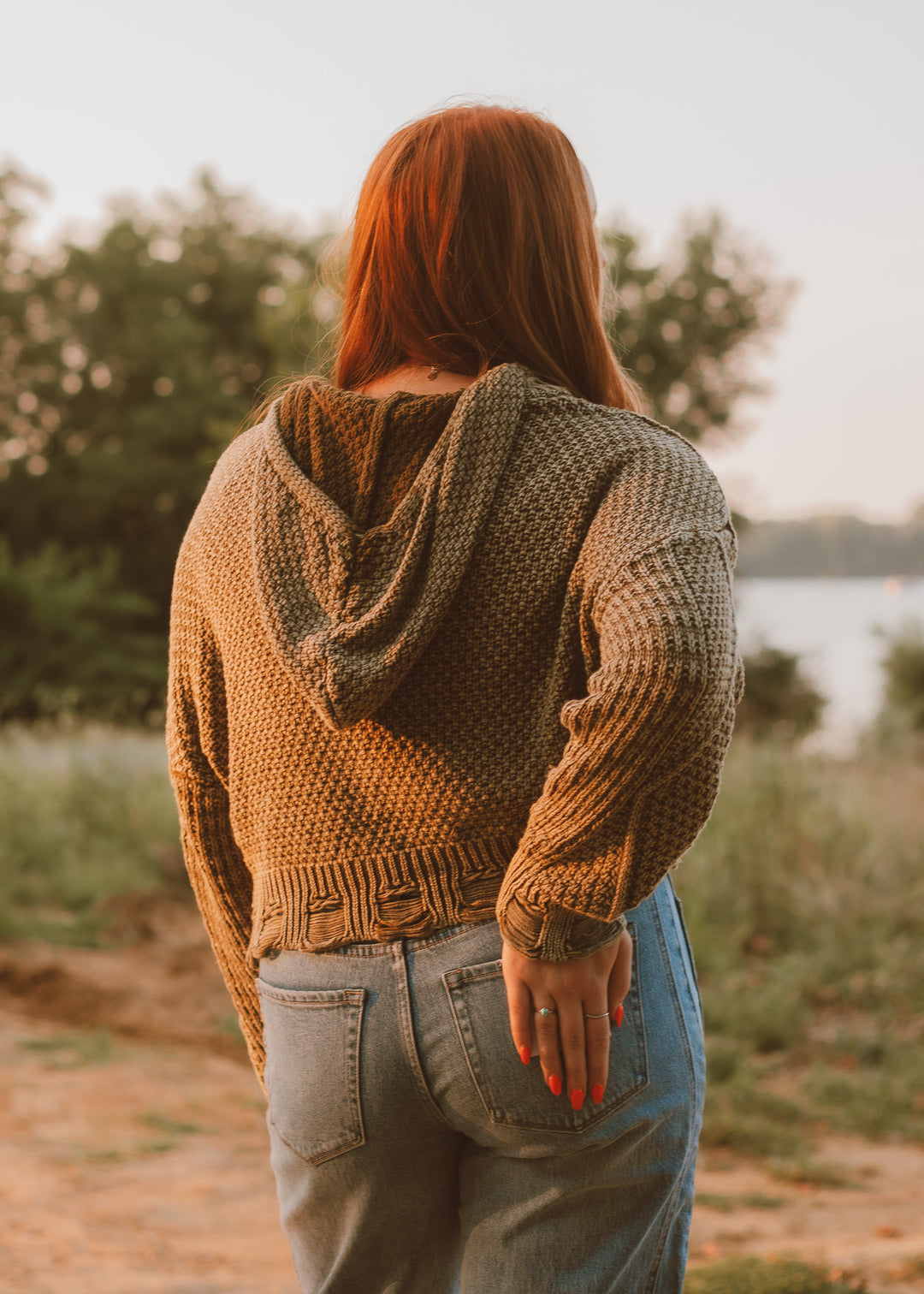 Person wearing a green knitted sweater and jeans standing outdoors by a body of water.