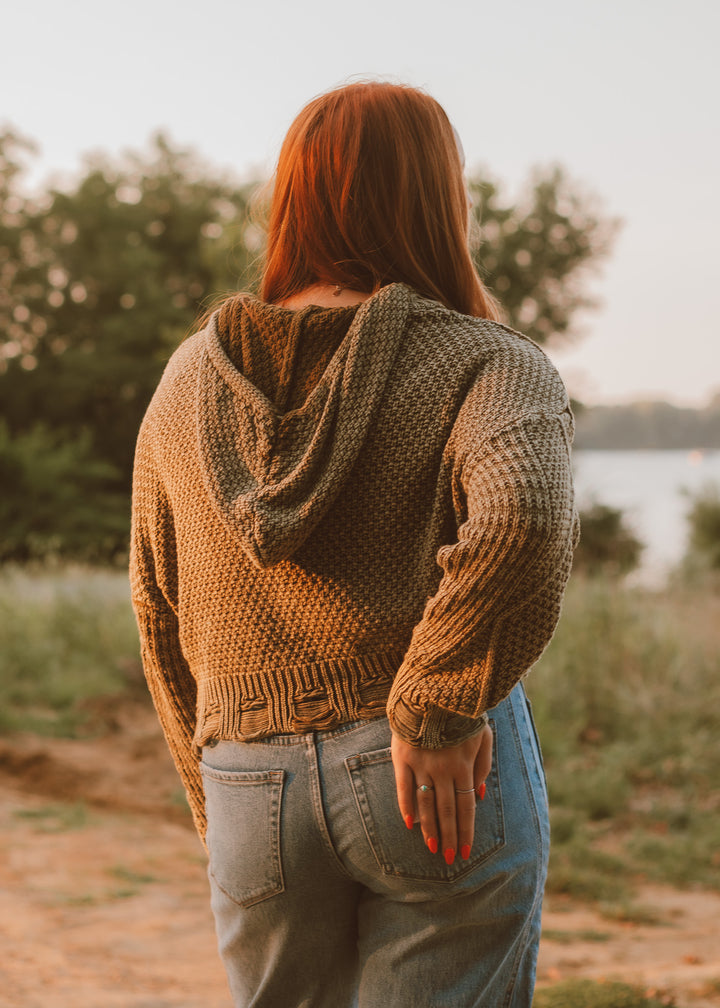 Person wearing a green knitted sweater and jeans standing outdoors by a body of water.