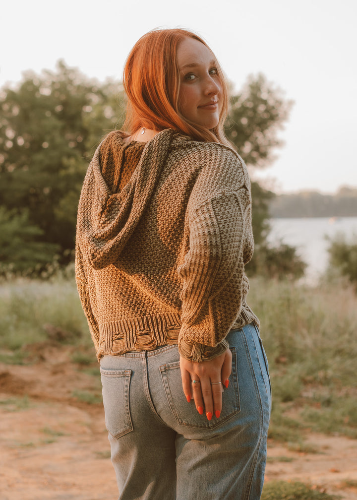 Woman wearing a green knitted hoodie and blue jeans standing outdoors with trees and a lake in the background.