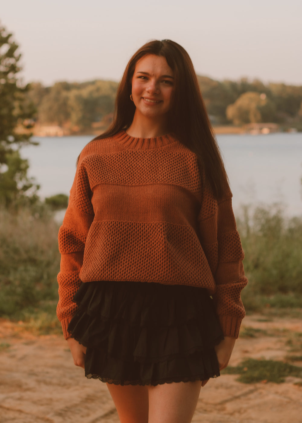 Woman wearing a brown sweater and black skirt standing by a lake.