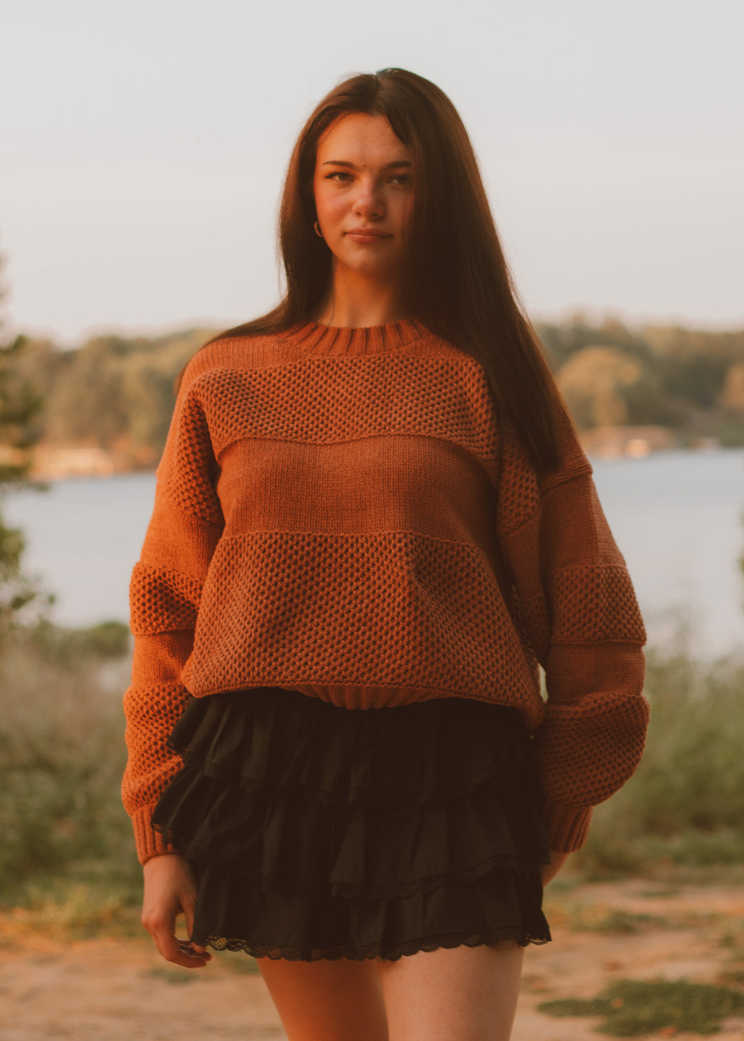 Woman wearing a brown knitted sweater and black skirt standing by a lake.
