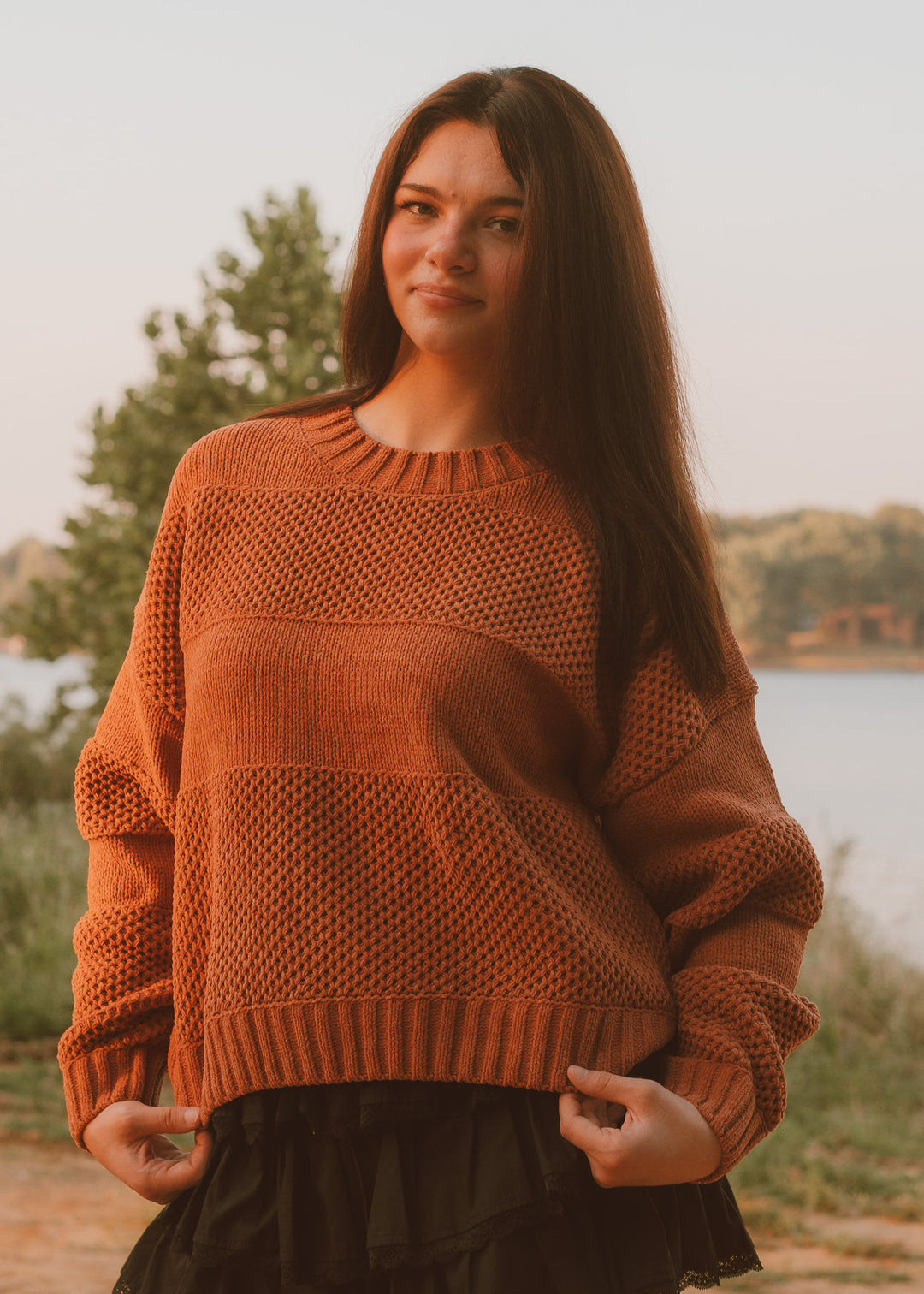 Woman wearing a brown sweater standing outdoors by a lake.