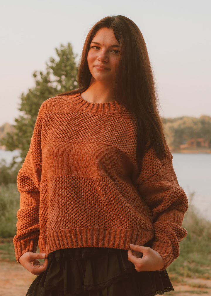 Woman wearing a brown sweater standing outdoors by a lake.