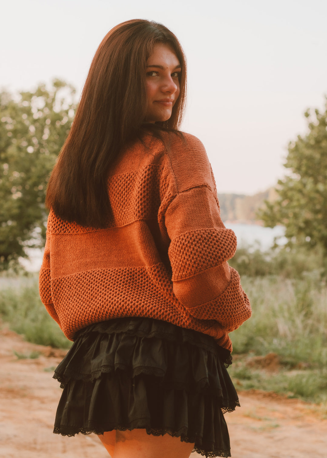 Woman wearing an orange sweater and black skirt standing outdoors with trees and a lake in the background
