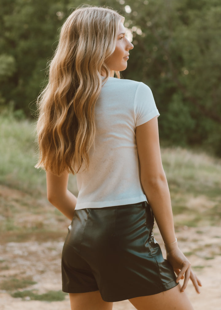 Woman with long blonde hair wearing a white t-shirt and dark shorts standing in a natural setting.