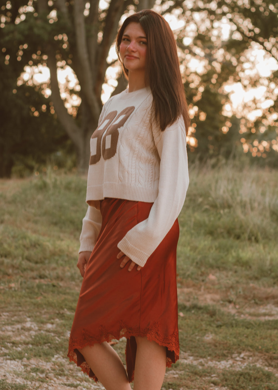 Woman wearing a white sweater with brown design and orange skirt standing in a field.