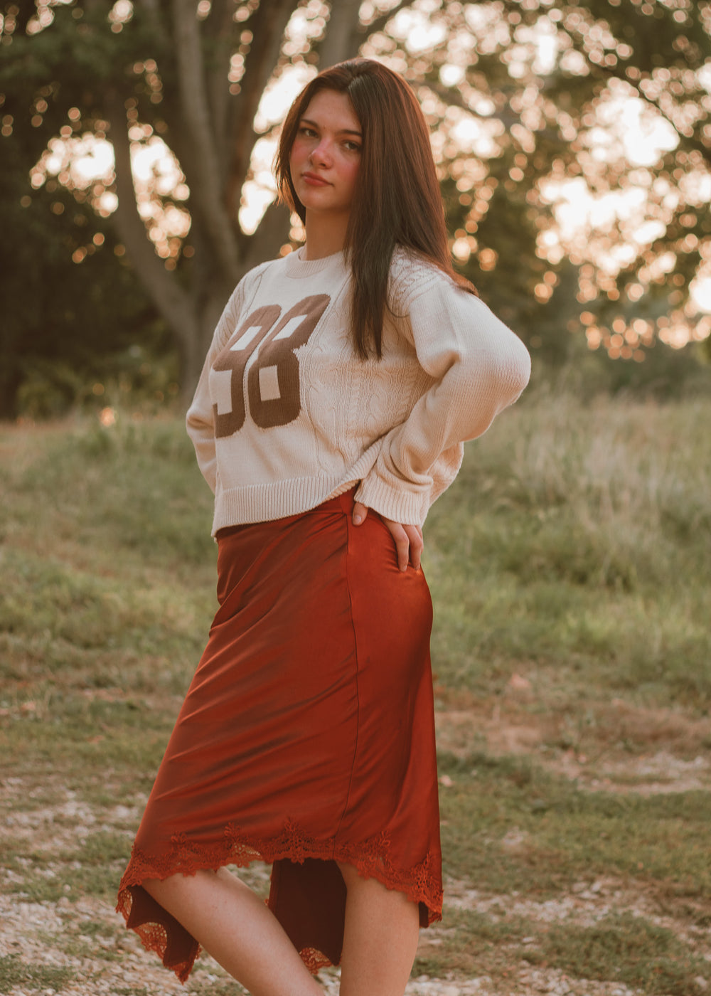 Woman wearing a beige sweater with large brown numbers and a red skirt standing in a field.