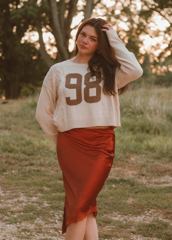Woman wearing a white sweater with the number 98 and a burnt orange skirt standing in a field.