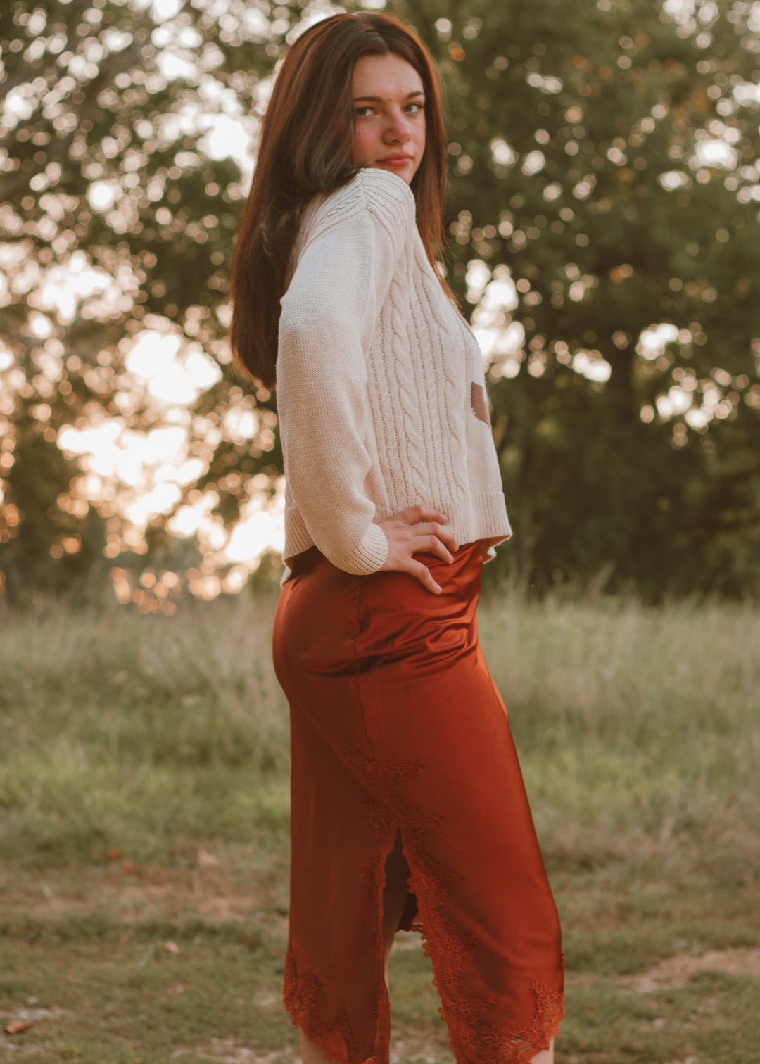 Woman in a white sweater and orange skirt standing in a field with trees in the background