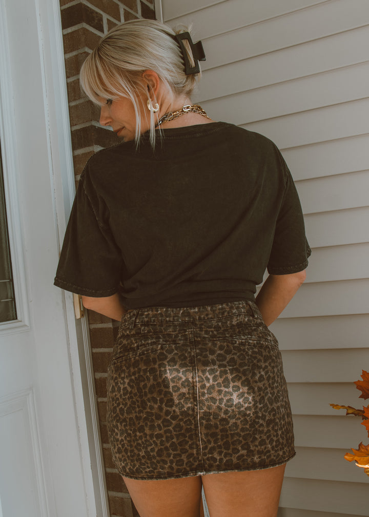 Person wearing a black t-shirt and leopard print skirt standing outside a house.