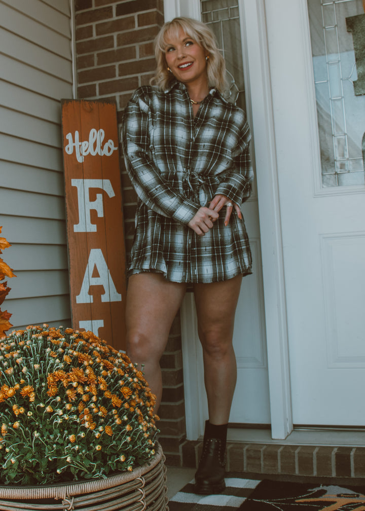 Woman in plaid dress standing in front of a 'Hello Fall' sign with pumpkins.