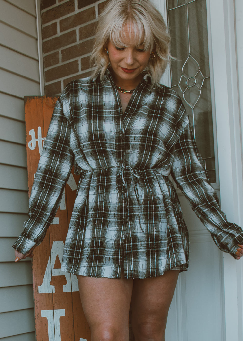 Woman wearing a plaid dress standing in front of a door with a wooden sign.