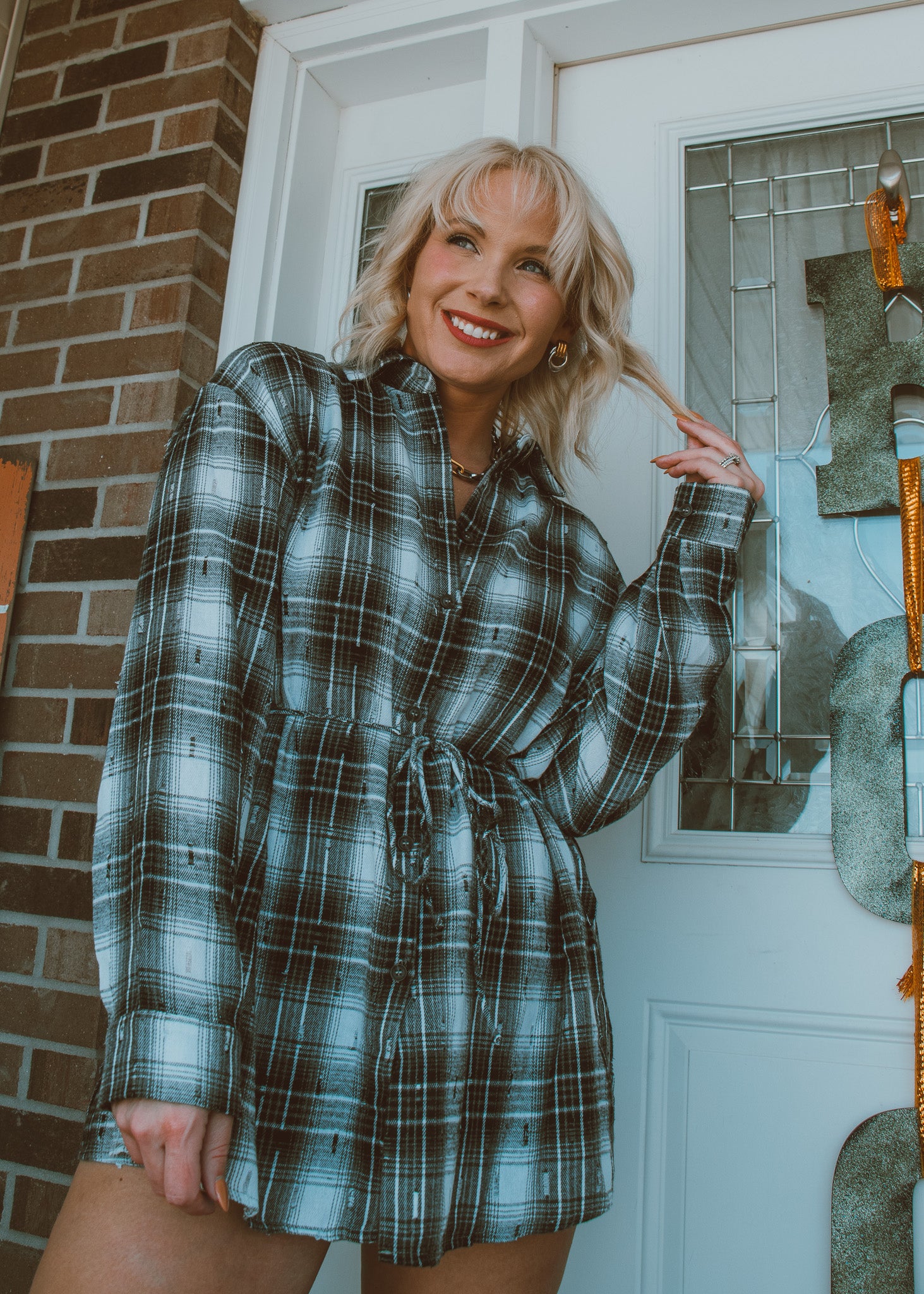 Woman wearing a plaid romper standing in front of a door with a brick wall background