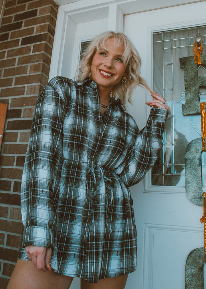 Woman wearing a plaid romper standing in front of a door with a brick wall background