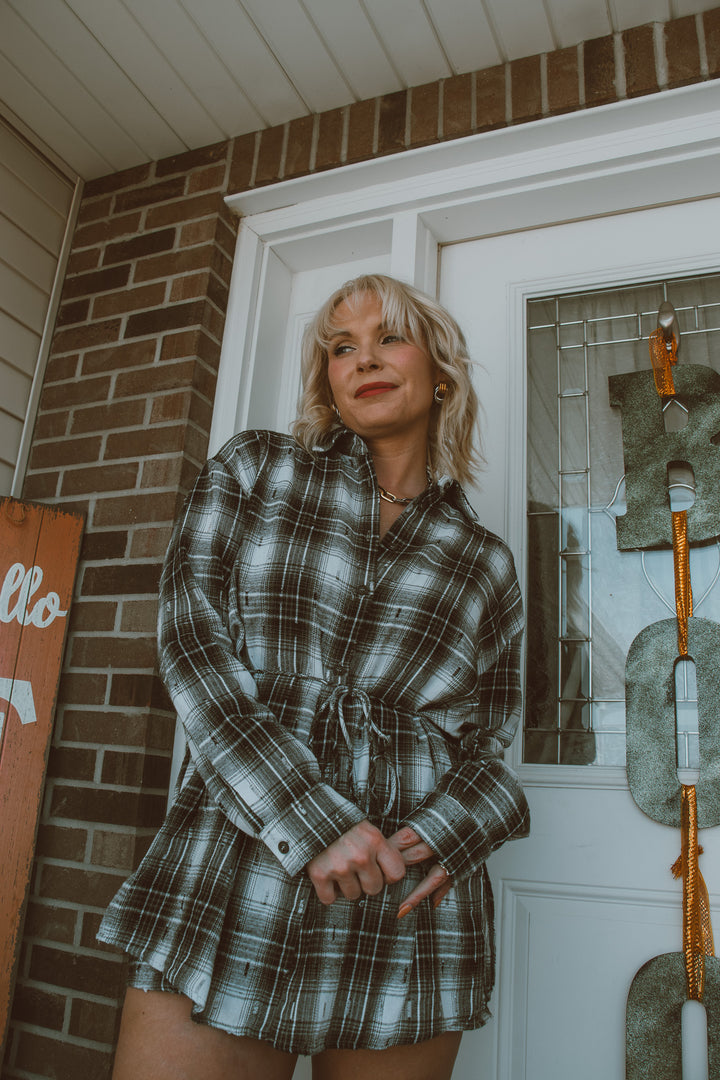 Woman in a plaid romper standing on a porch with a brick wall and door in the background.