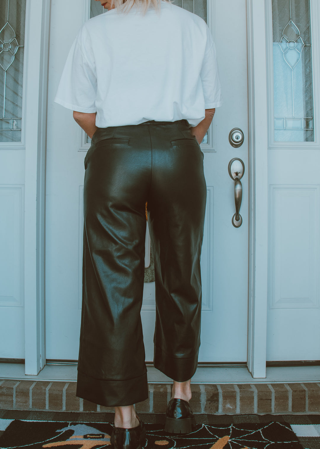 Person wearing black leather pants and a white shirt standing in front of a door.