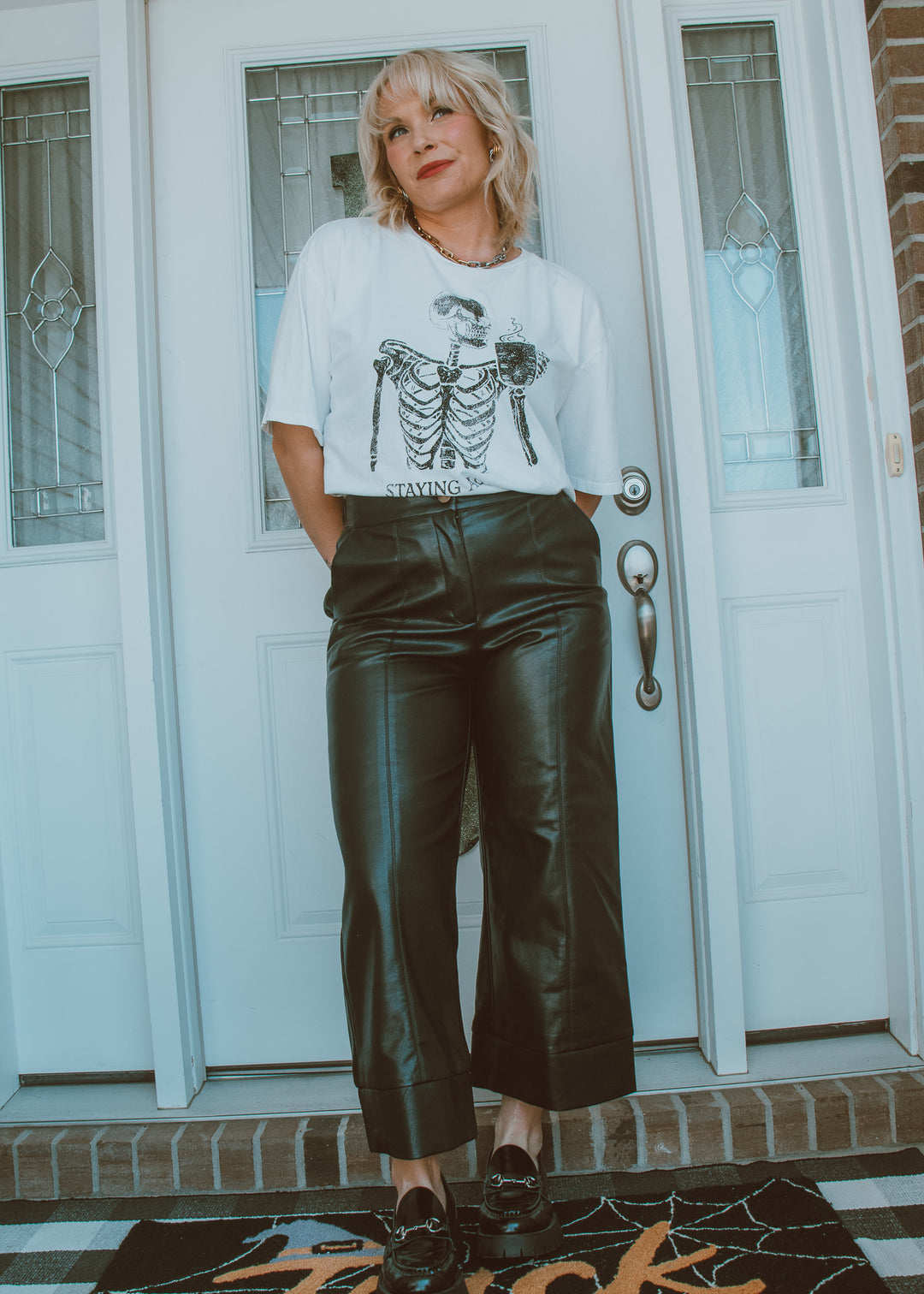 Person wearing a white t-shirt with a skeleton design and black leather pants standing in front of a door.