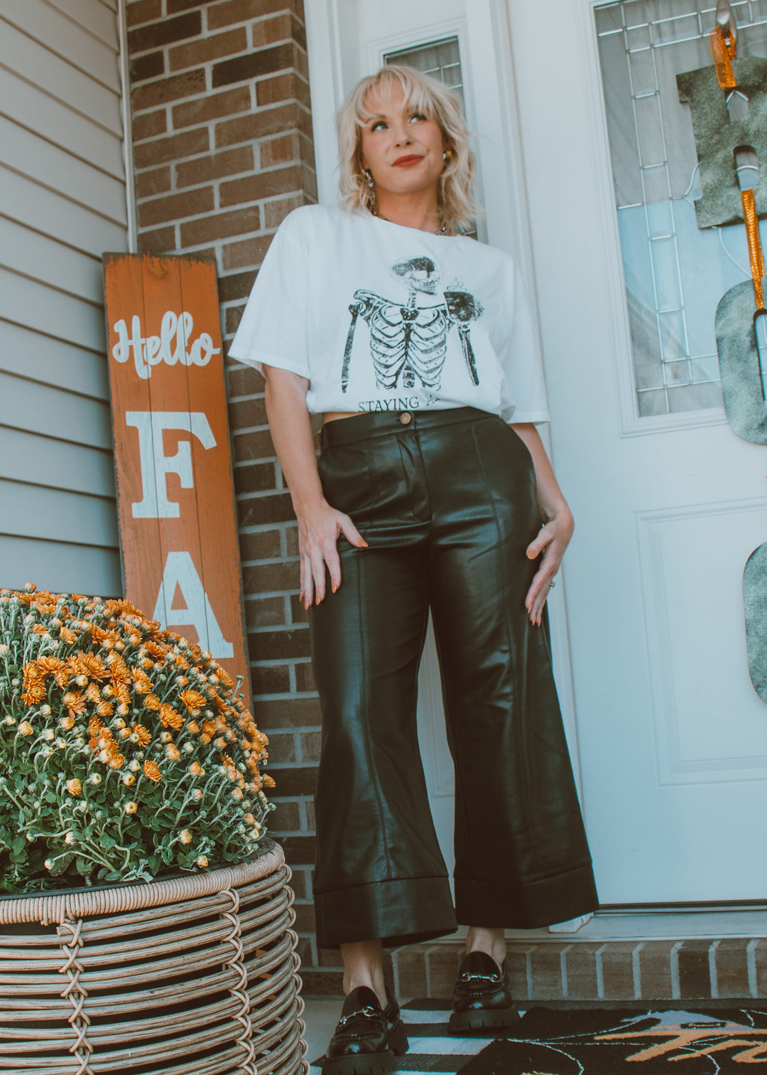 Woman wearing a white t-shirt with a skeleton design and black pants standing outside a house.