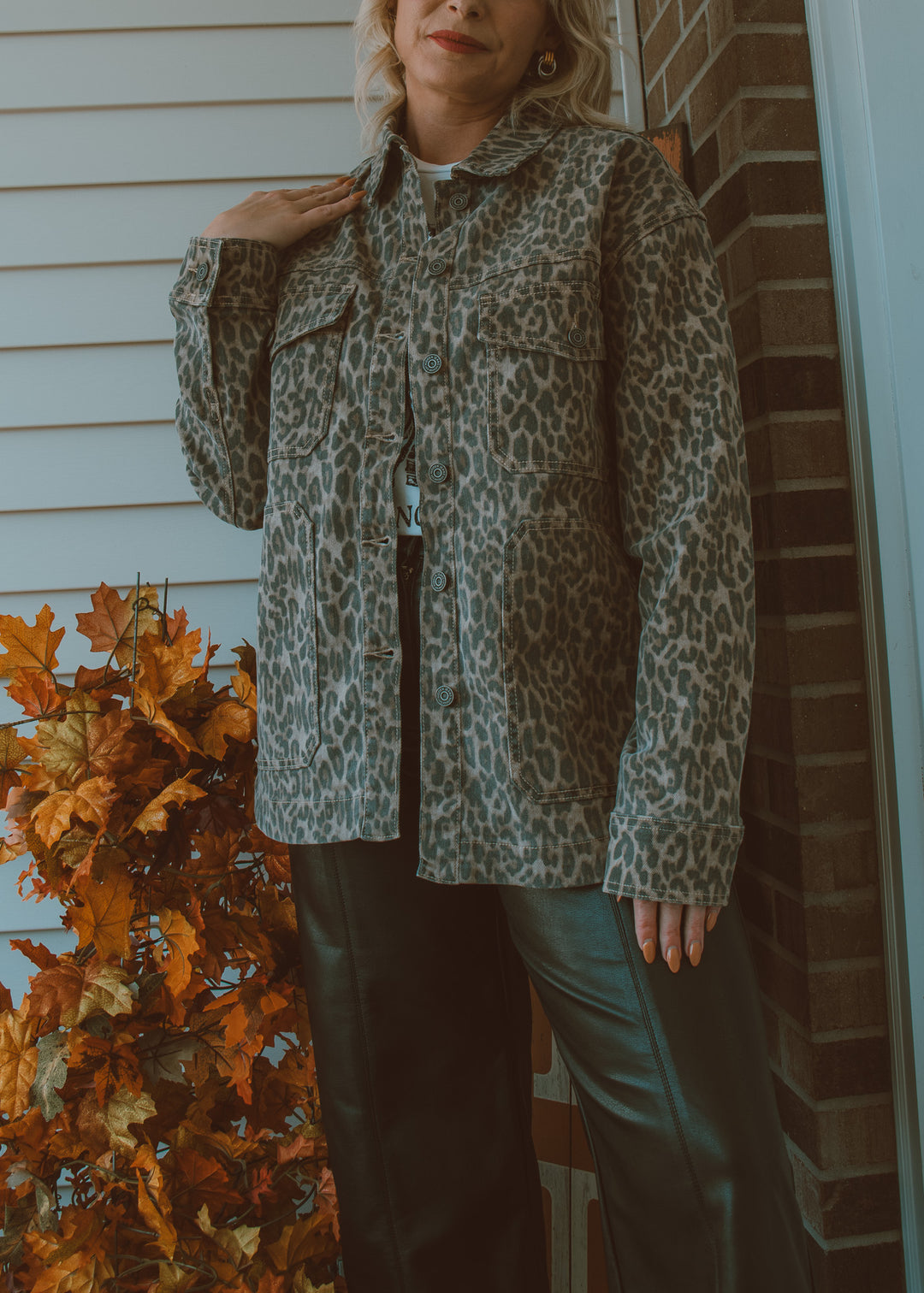 Person wearing a leopard print jacket standing in front of a brick wall with autumn leaves.