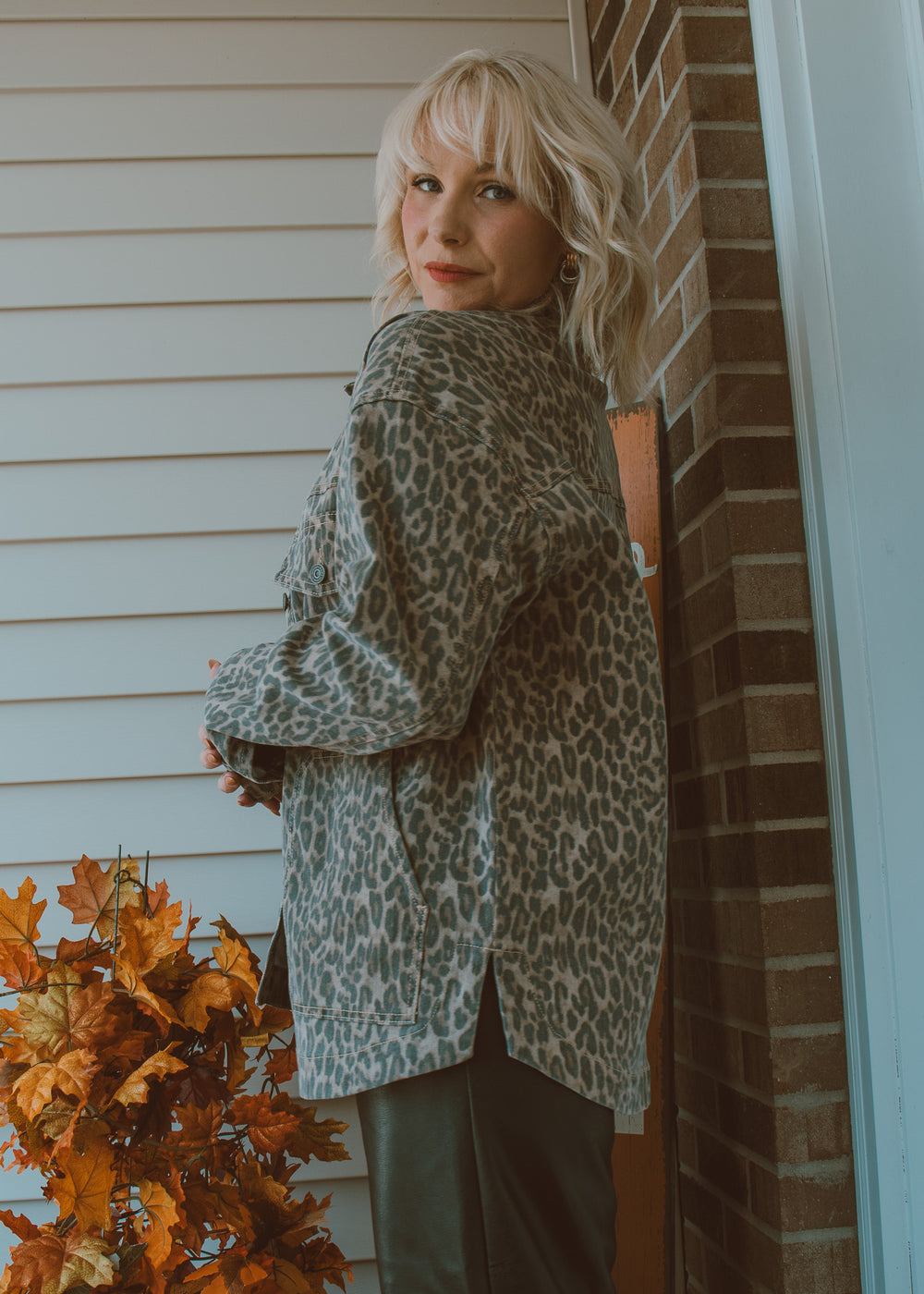 Woman wearing a leopard print jacket standing against a brick wall with autumn leaves.