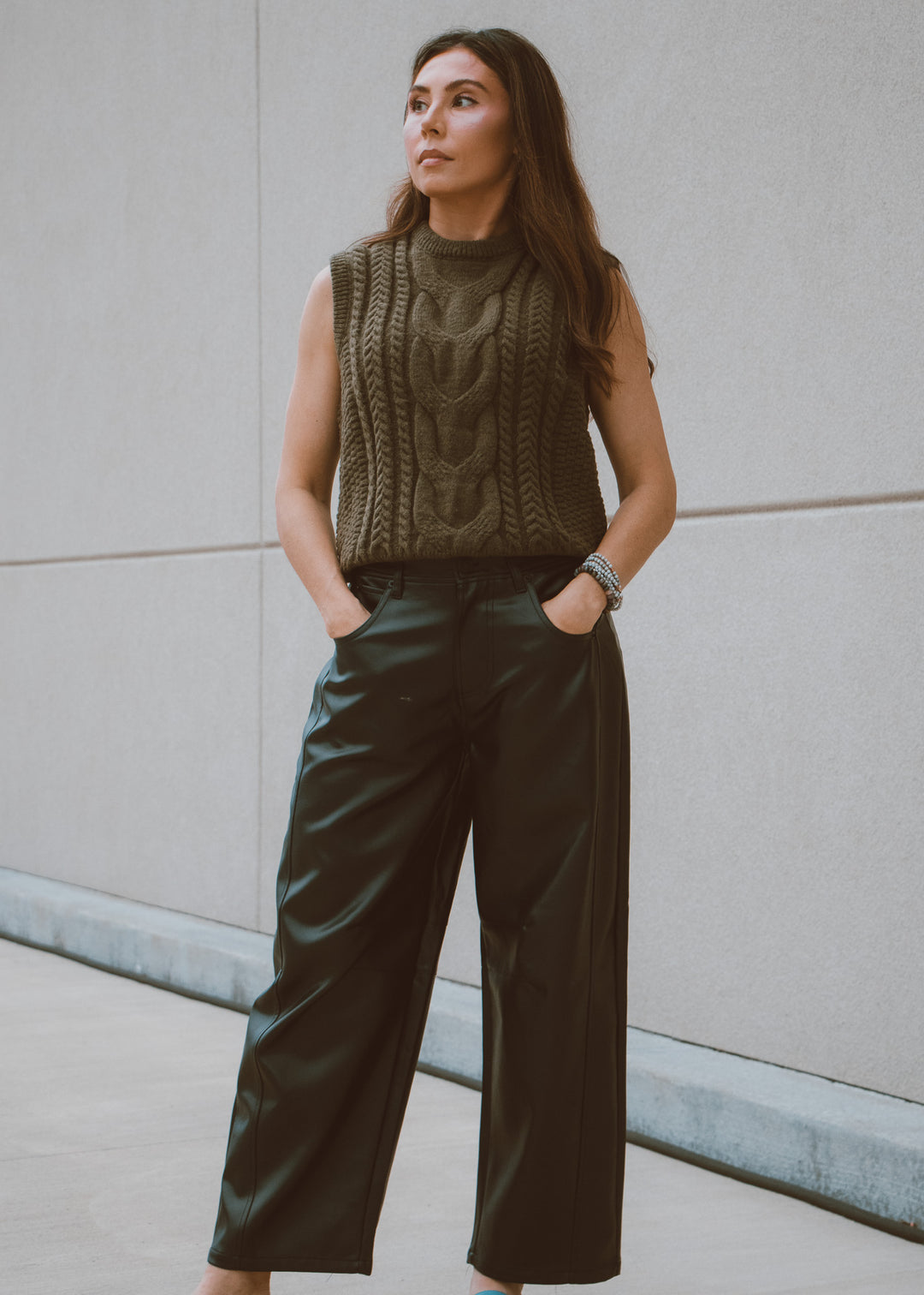 Woman wearing a green knitted sleeveless sweater and leather pants against a plain wall.