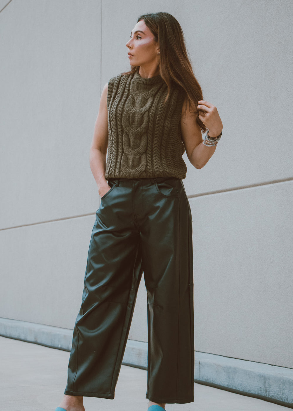 Woman wearing a green knitted sleeveless sweater vest and wide-leg  leather pants against a gray wall.