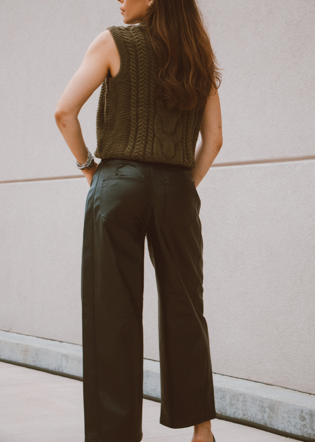 Woman wearing a green knitted sleeveless sweater and wide-leg leather pants against a light-colored wall.