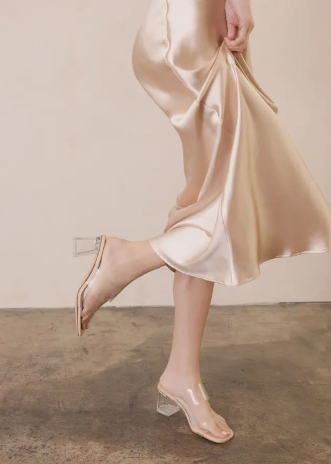 Person wearing a beige satin skirt and clear high-heeled shoes on a neutral background